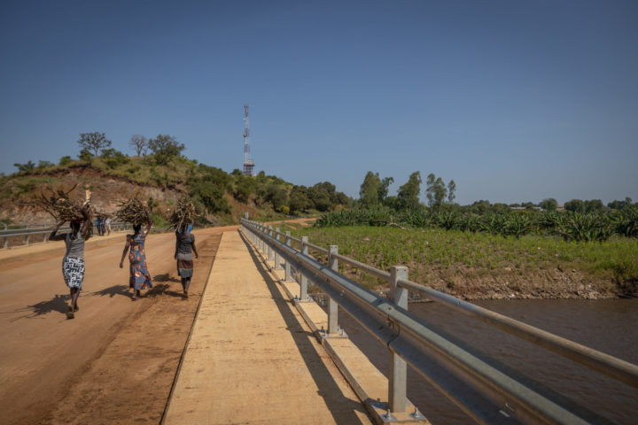 Kolme naista kävelee siltaa joen yli, kantaen polttopuita päidensä päällä.