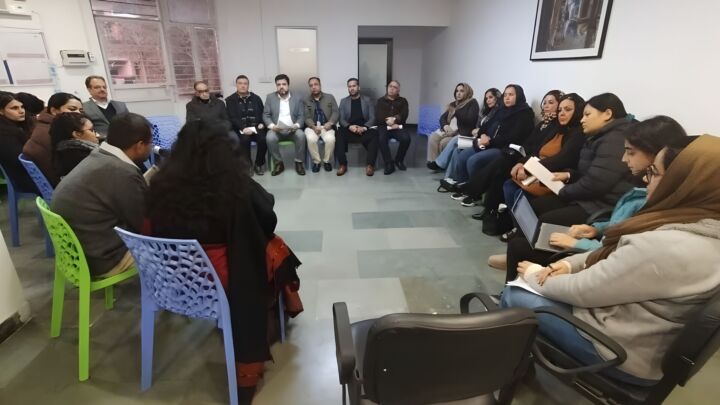 The picture is from an awareness session about legal pathways for refugees. There's many people sitting in a circle listening to someone talk.