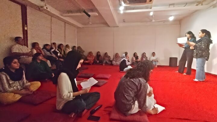 The photo is from an awareness session about legal pathways for refugees. Older people are sitting on chairs and younger people on the floor. They are listening to two people at the front reading something.