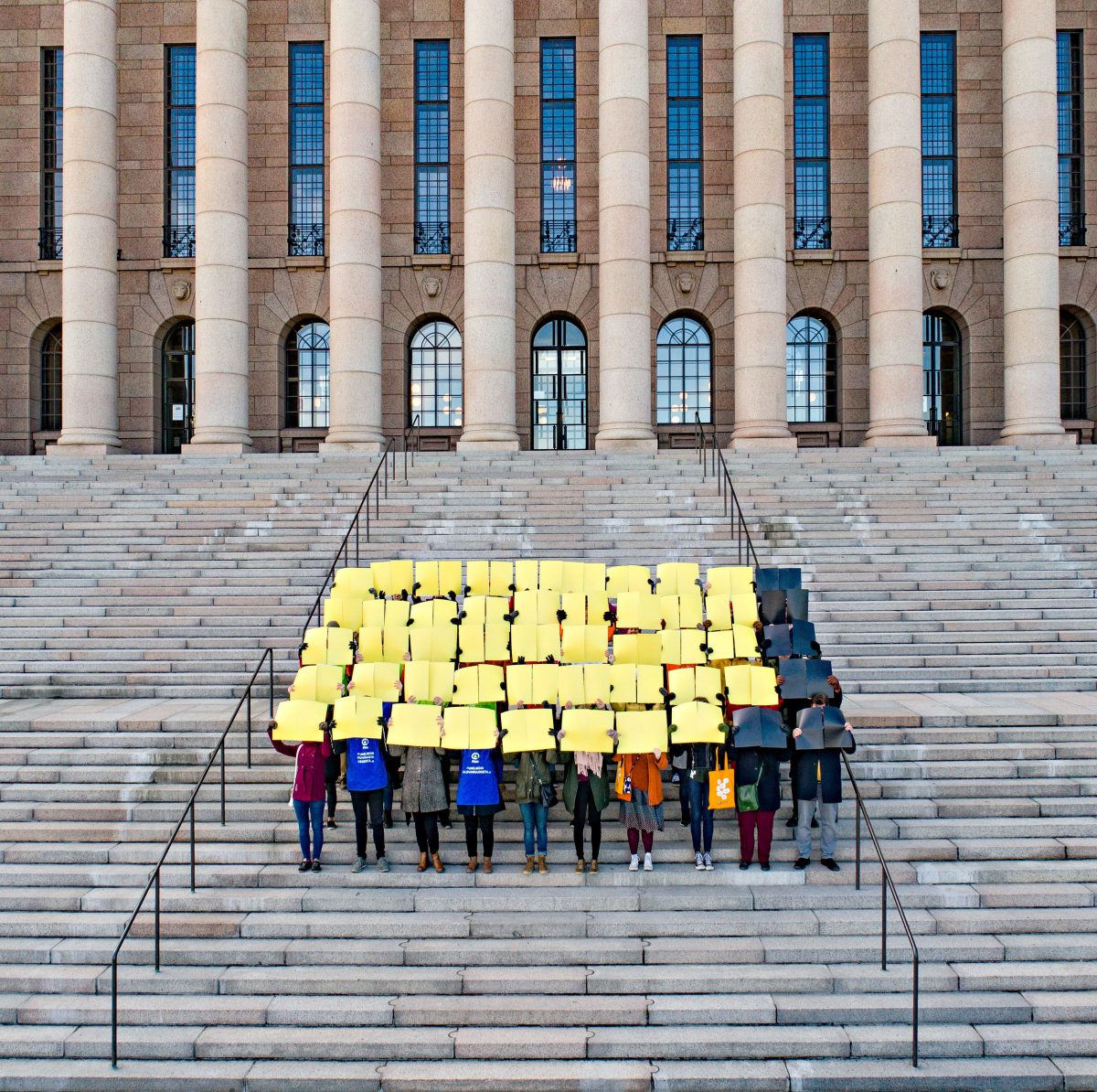 Ihmisiä eduskuntatalon portailla pitämässä kylttejä joista suurin osa on keltaisia
