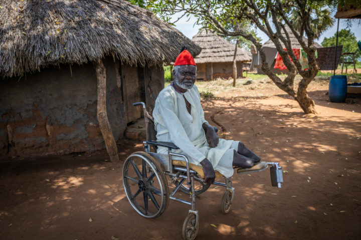 Man sitting in a wheel chair. In the background houses with thatched roofs.