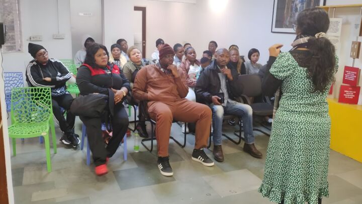 The photo is from an awareness session about legal pathways for refugees. A small group of adults are sitting on chairs and listening to a woman speak.