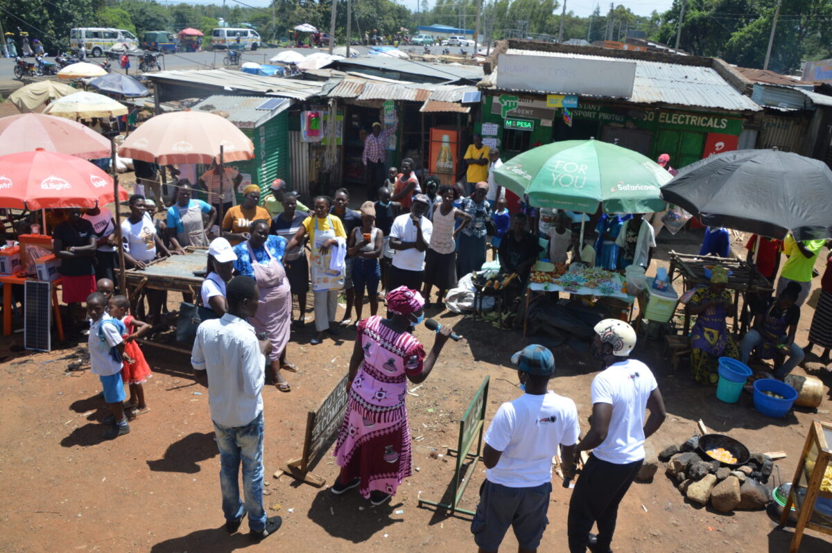 People gathering to listen to a woman talking into a microphone. In the background market stalls.