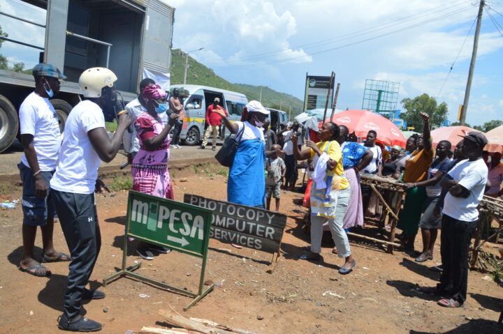 People gathering alongside a road, some of them speaking in a microphone and some of them listening. In the background market stalls and trucks.