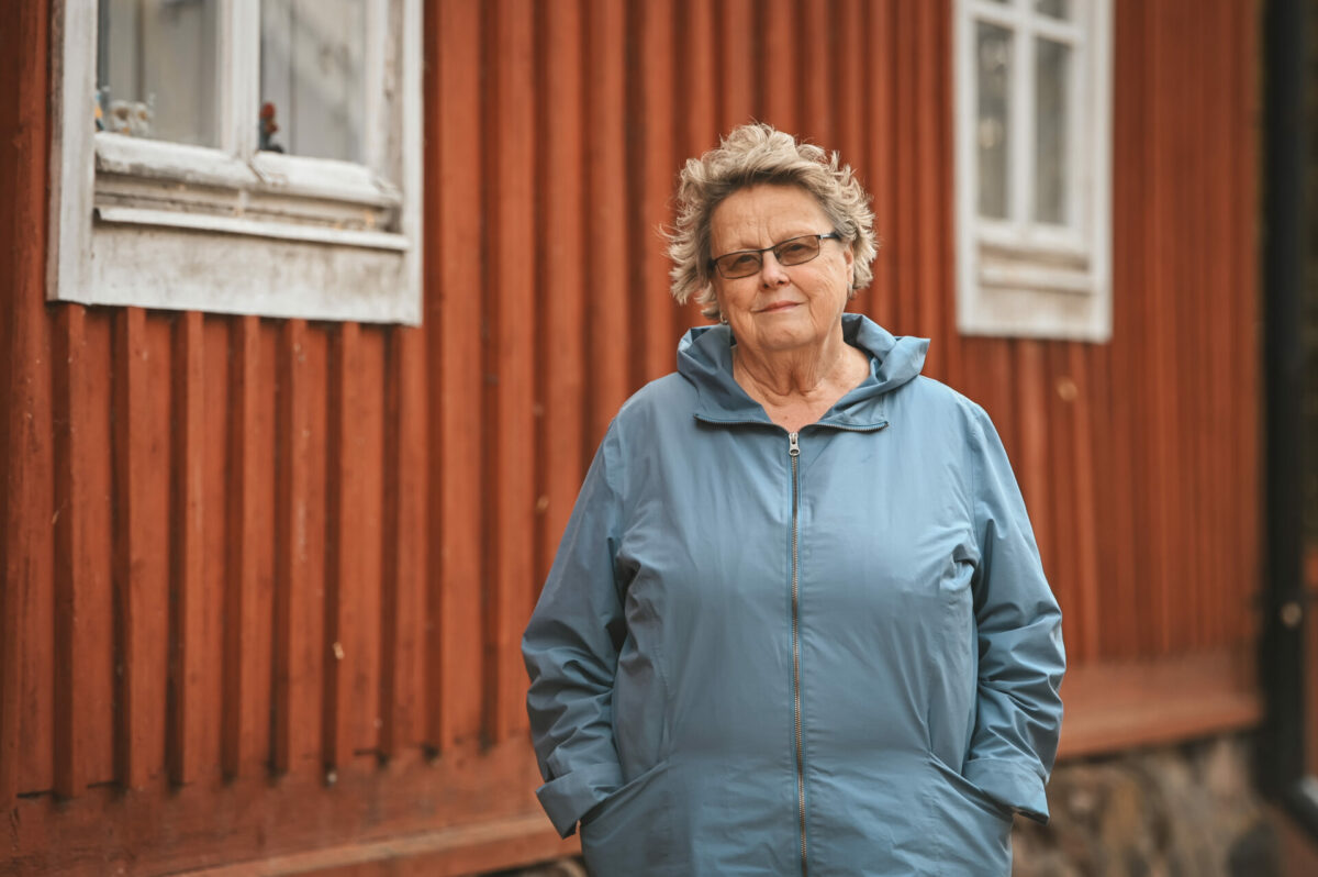 Helena Ranta seisoo sininen takki yllään punamultaisen talon edessä.