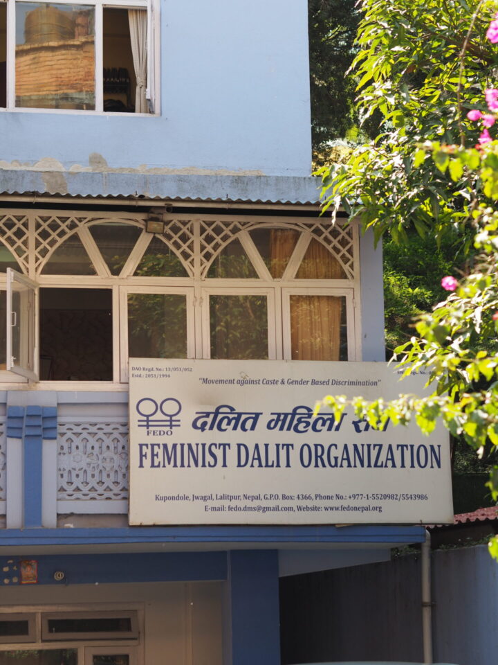 Kuvassa näkyy FEDO:n toimiston etuosa, rakennuksen seinät ovat vaaleansiniset ja rakennukseen on kiinnitetty kyltti jossa lukee valkoisella pohjalla, tummansinisin kirjaimin "Feminist Dalit Organization". Rakennuksen oikealla puolella kasvaa ja kukkii puu. 