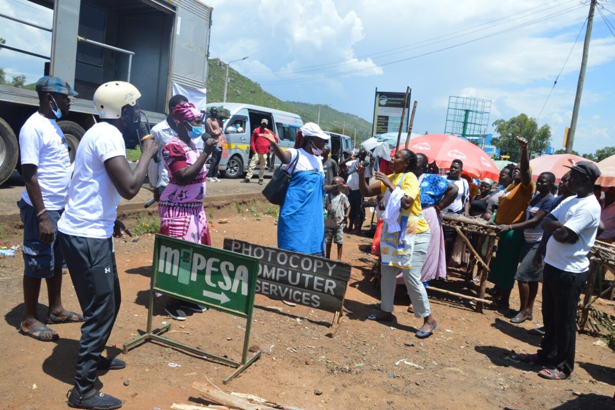 People gathering alongside a road, some of them speaking in a microphone and some of them listening. In the background market stalls and trucks.