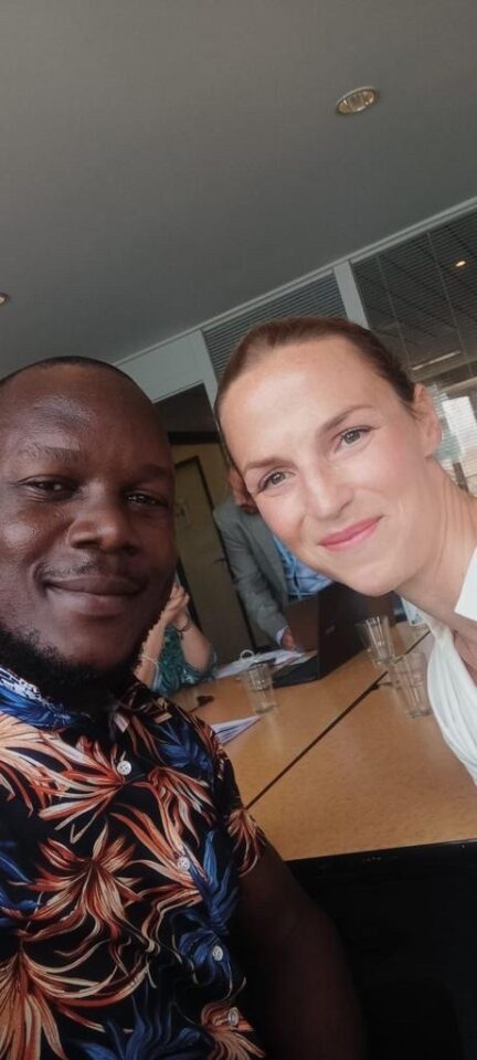 Kuvassa kaksi ihmistä katsoo suoraan kameraan. Vasemmalla tummaihoinen mies (Ishmael Bahati) ja oikealla vaalea nainen (Elina Vuola). Taustalla kokoushuone.