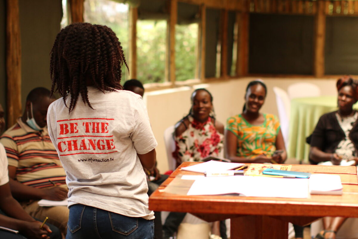 A woman standing with her back to the camera, with the text "Be the Change" on her back, and other women sitting behind a table listening to her.