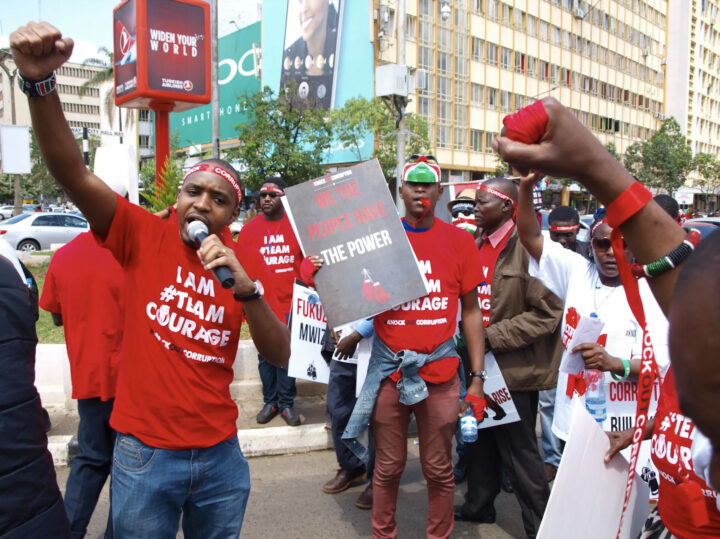 Kenialaisia mielenosoittajia kadulla. Mielenosoittajilla on punaiset t-paidat, joissa lukee ”I AM #TEAM COURAGE” ja pään ympärillä punaiset nauhat, joissa lukee ”Knock out corruption”. Toisessa reunassa seisova mies huutaa mikrofoniin ja pitää kättä nyrkissä. Keskellä kuvaa mies viheltää pilliin ja pitelee kylttiä, jossa lukee ”WE THE PEOPLE HAVE THE POWER”.