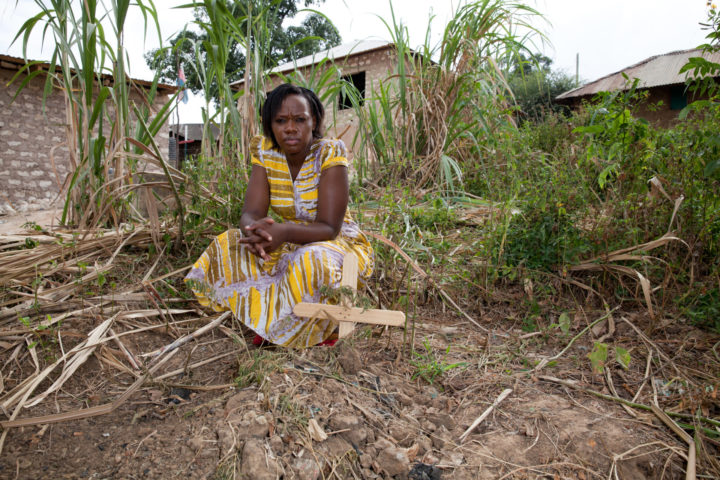 Woman in yellow dress next to a wooden cross standing in the mud, surrounded by grass and sticks. In the background small brick houses.