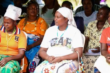 Participants of an event looking to stage, they are visibly listening to someone speak. Audience is made of Kenyan women, some are wearing traditional dressess and some have their hair covered.