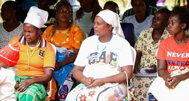 Participants of an event looking to stage, they are visibly listening to someone speak. Audience is made of Kenyan women, some are wearing traditional dressess and some have their hair covered.