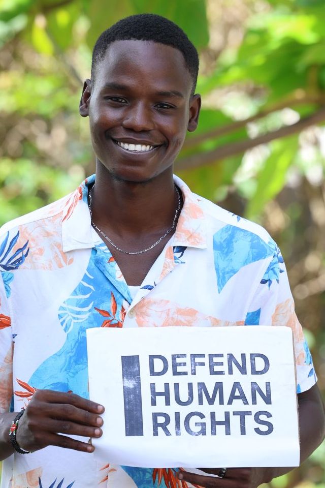 Mustapha Aza's brave action. They are looking into the camera smiling and holding a sign that says "I Defend Human Rights."