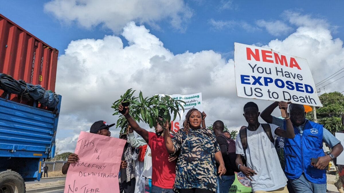 Phyllis and a group of demonstrators walking on a road. One demonstrator holds a sign saying "NEMA you have exposed us to death".