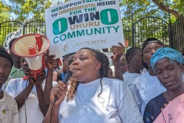 Lähikuvassa Phyllis Omido puhumassa mielenosoittajille oikeustalon edessä. Taustalla kyltti, jossa lukee "We support access to environmental justice, Owinouhuru community" sekä megafoni. 
