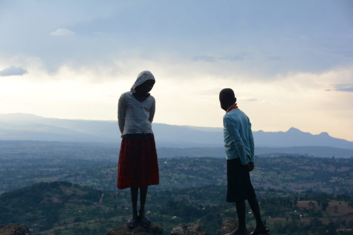 A young girl and a boy watching down the valley on a cliff. Dark clouds above them. In the background hills and a bright sky.